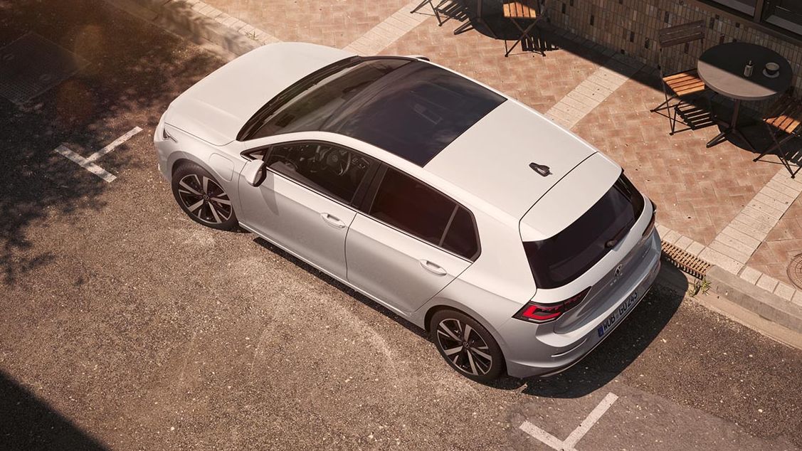 Volkswagen Golf je parkiran ob pločniku v mestnem okolju, viden od zgoraj pod kotom. Ima panoramsko stekleno streho, temnejša stekla zadaj in športna platišča. Ob robu so mize in stoli kavarne na tlakovanem pločniku.
