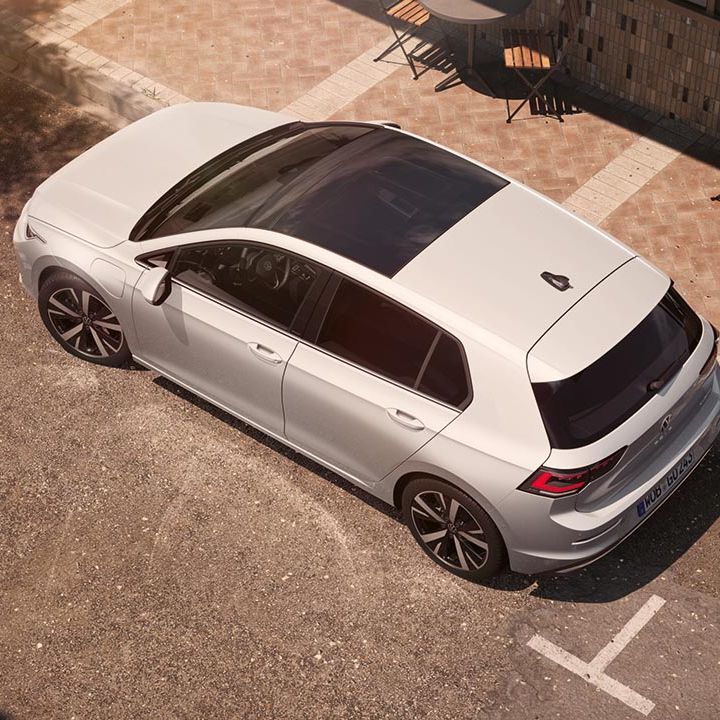 Volkswagen Golf je parkiran ob pločniku v mestnem okolju, viden od zgoraj pod kotom. Ima panoramsko stekleno streho, temnejša stekla zadaj in športna platišča. Ob robu so mize in stoli kavarne na tlakovanem pločniku.