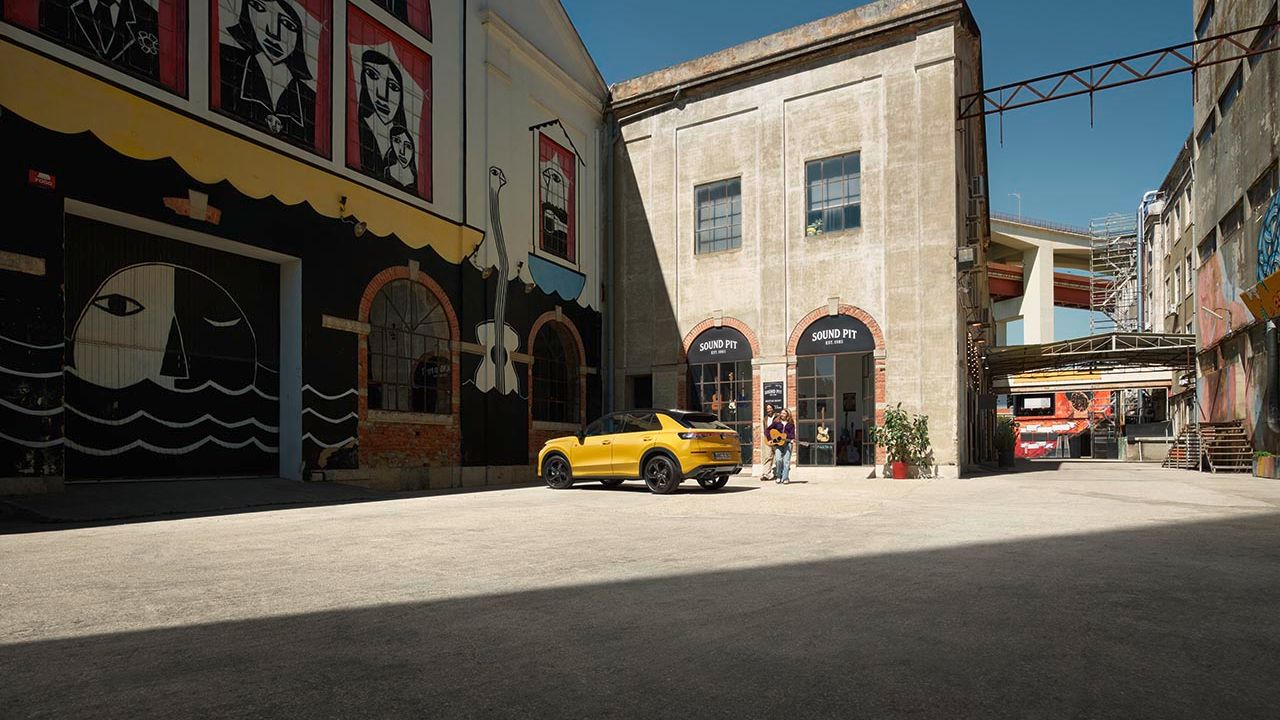 Rumeno športno vozilo parkirano na praznem dvorišču, obdanem z industrijskimi stavbami in uličnimi grafitami. Dva človeka stojita ob vozilu.