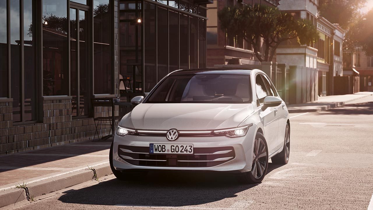 Bel avtomobil Volkswagen Golf stoji ob robu sončne mestne ulice, obrnjen rahlo proti kameri. Stavbe z veliko stekla in drevesa v ozadju nakazujejo mirno urbano okolje. Svetloba poznega popoldneva ustvarja mehke sence na cesti in karoseriji.