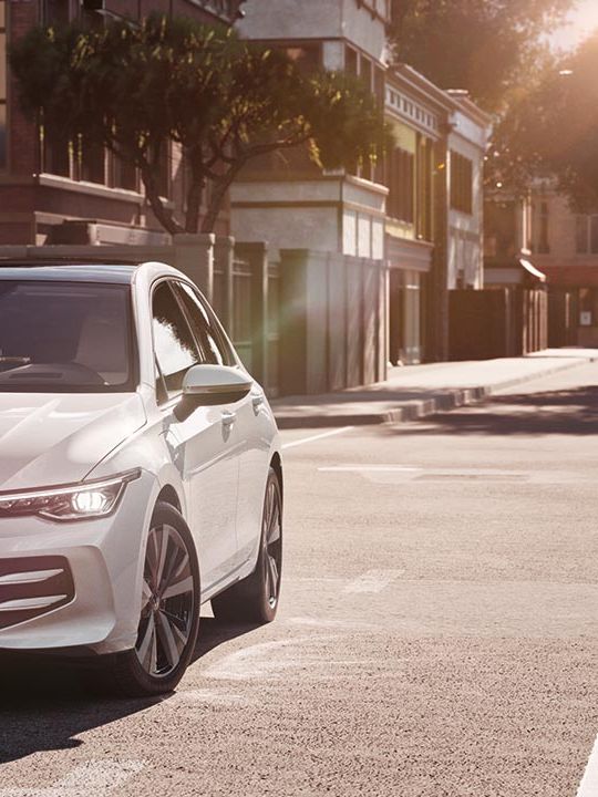 Bel avtomobil Volkswagen Golf stoji ob robu sončne mestne ulice, obrnjen rahlo proti kameri. Stavbe z veliko stekla in drevesa v ozadju nakazujejo mirno urbano okolje. Svetloba poznega popoldneva ustvarja mehke sence na cesti in karoseriji.