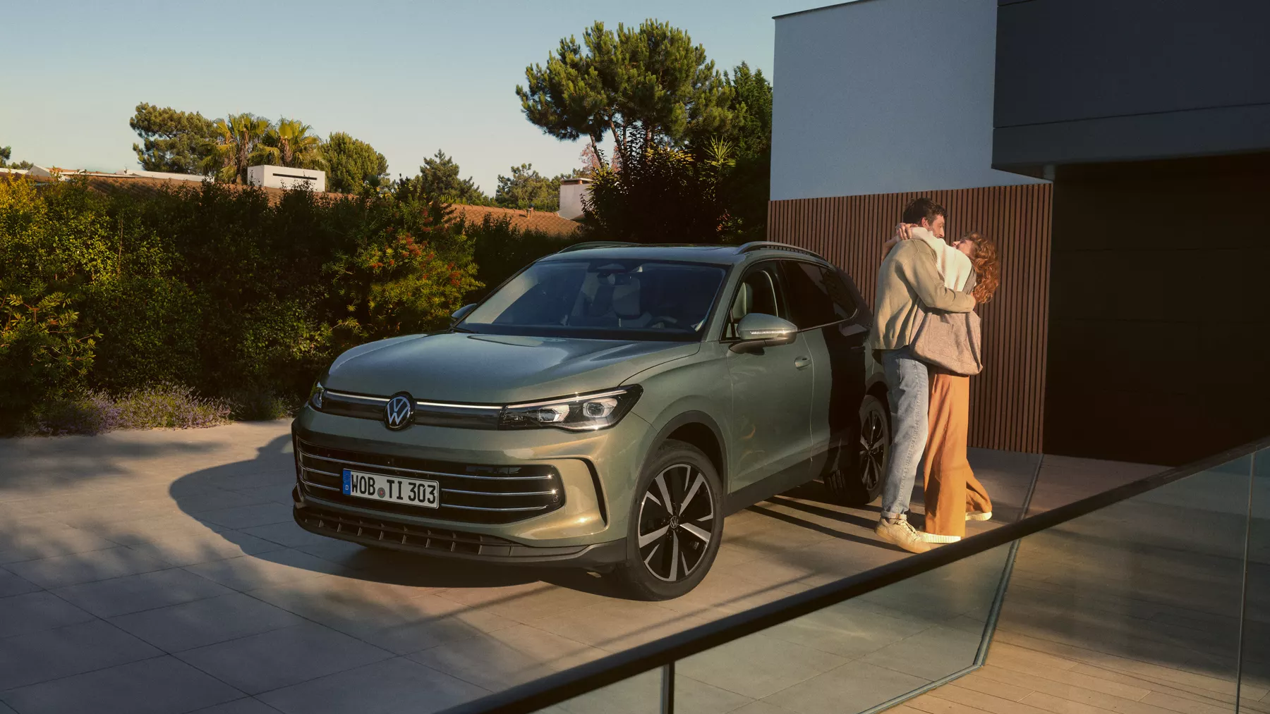 Pogled od spredaj na zelen VW Tiguan, parkiran na dovozu moderne stanovanjske hiše. Ob njem stoji mlad par in se objema.