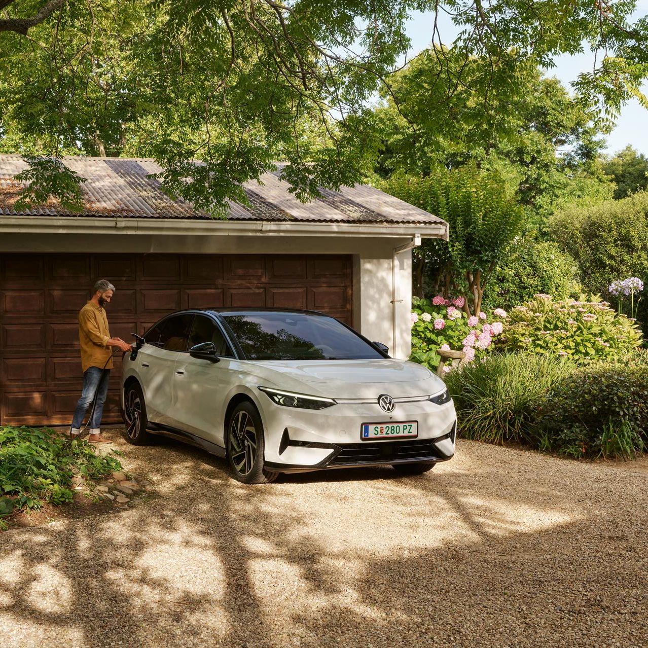 Moški parkira električni avto pred garažo na dvorišču obkroženem z zelenjem in cvetličnimi grmi.