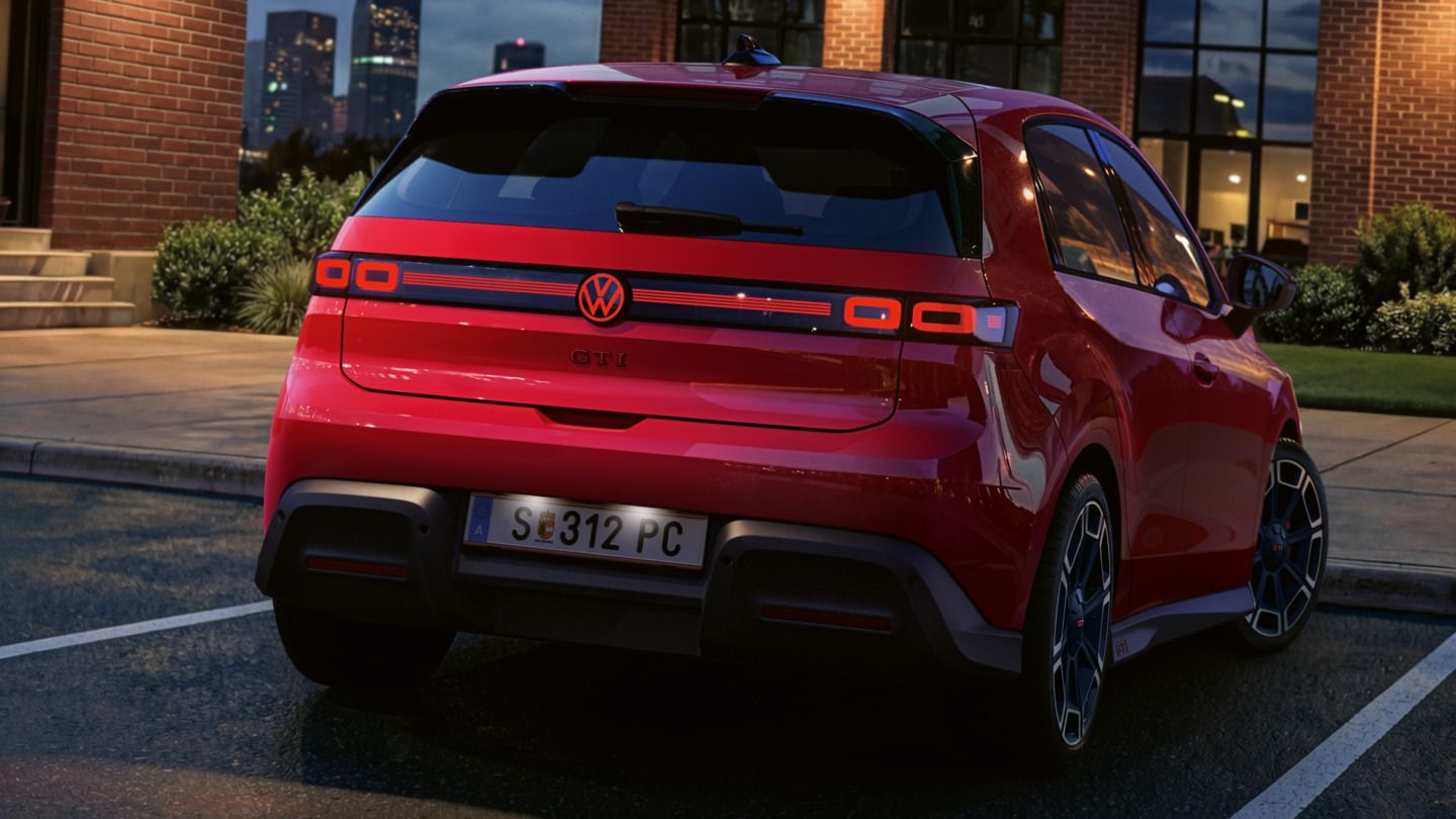 Pogled nazaj na rdeči VW ID. Polo GTI z vklopljenimi zadaj lučmi in osvetljenim logotipom, parkiran pred poslovno stavbo.