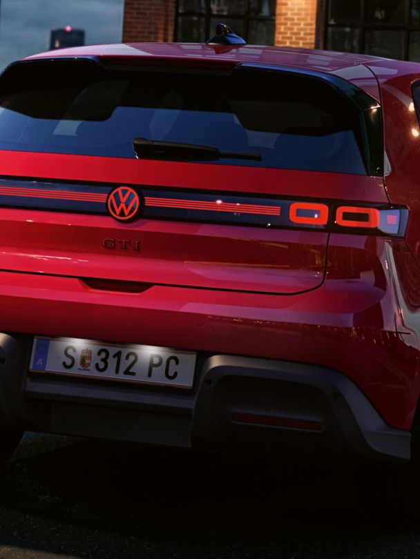 Pogled nazaj na rdeči VW ID. Polo GTI z vklopljenimi zadaj lučmi in osvetljenim logotipom, parkiran pred poslovno stavbo.