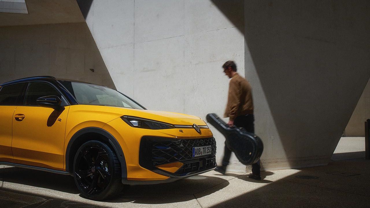 Moški s kovčkom za kitaro hodi mimo parkiranega rumenega VW T-Roc R-Line.