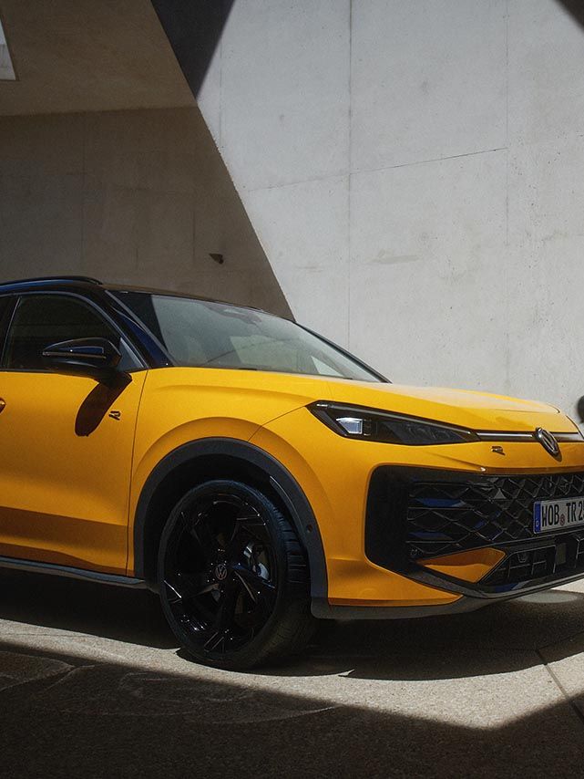 Moški s kovčkom za kitaro hodi mimo parkiranega rumenega VW T-Roc R-Line.