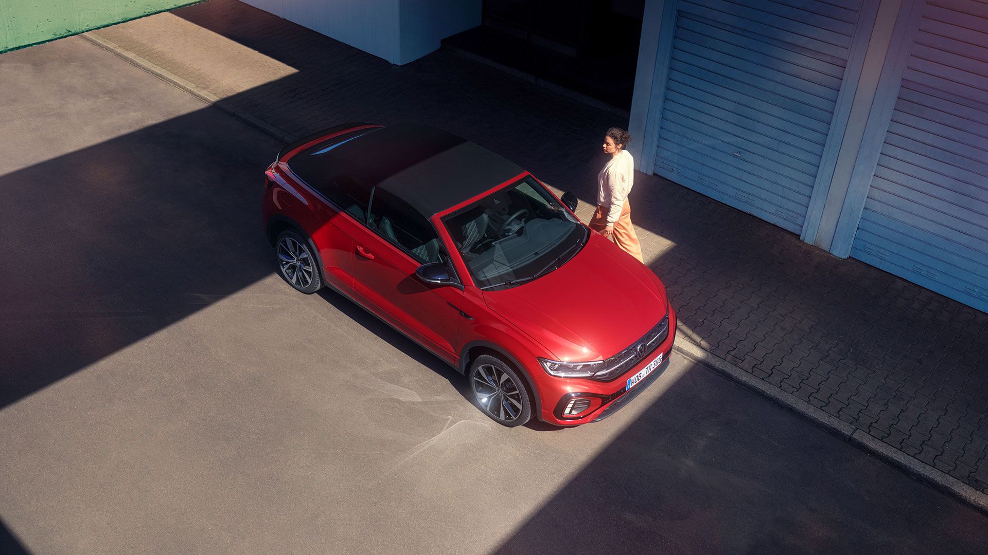 Rdeč VW T-Roc Cabriolet z odprto streho s ptičje perspektive. Parkiran je ob robu cestišča, mimo njega gre ženska. 