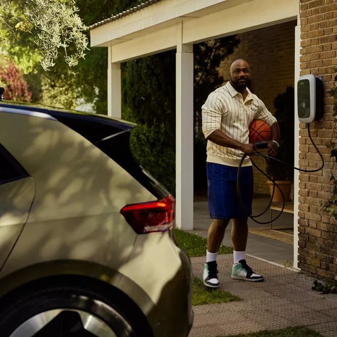 Oseba, ki drži košarkarsko žogo in kabel za polnjenje električnega vozila, stoji poleg hiše. Pred njim je parkirano vozilo. Okolica je zelena z drevesi in grmičevjem.
