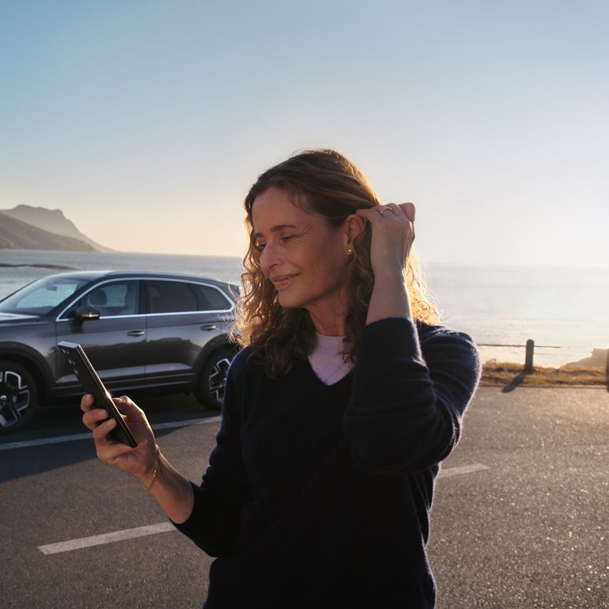 Pogled na nasmejano žensko z mobilnim telefonom v roki, morje in Touareg v ozadju.