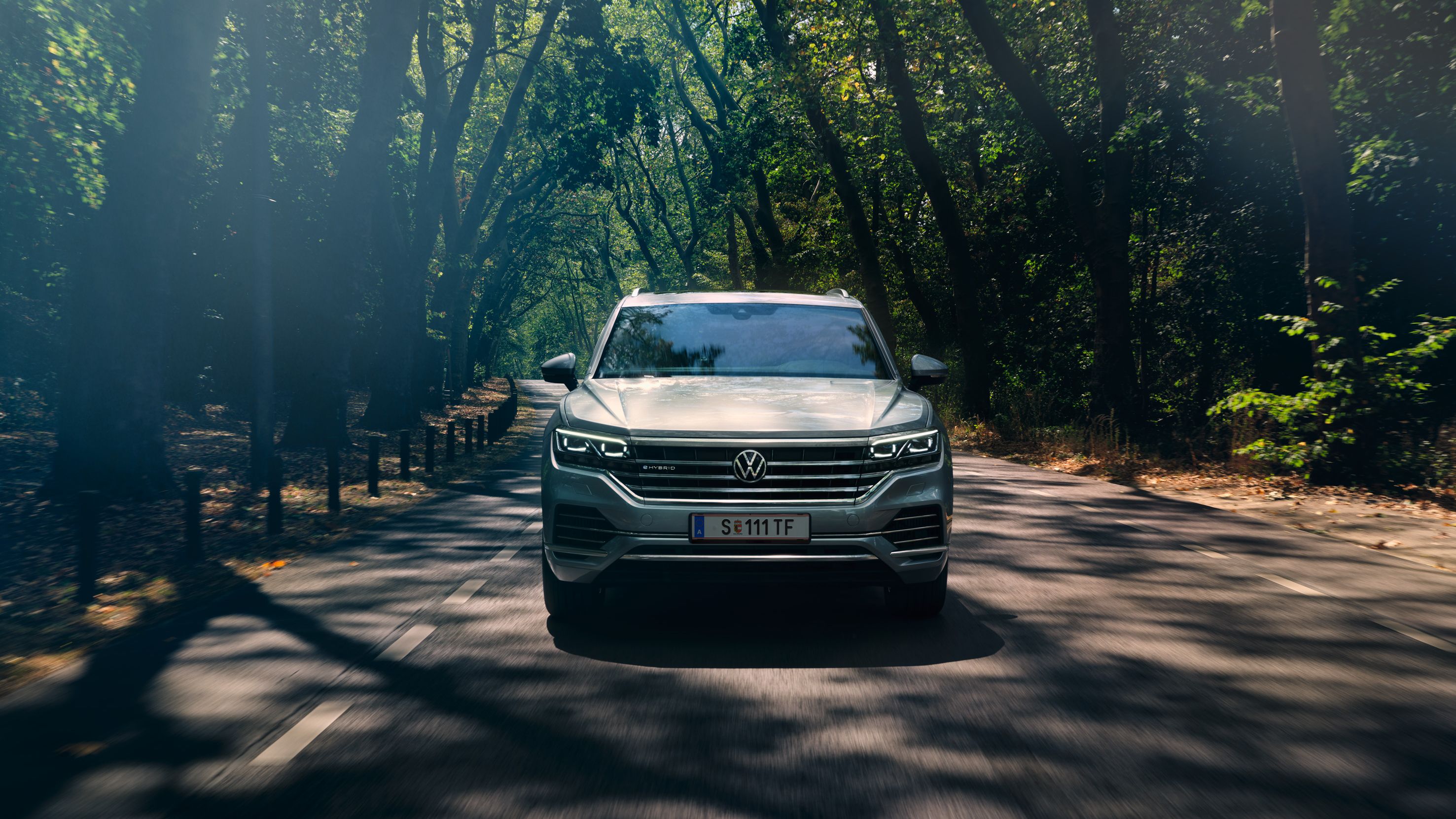 Volkswagen Touareg eHybrid srebrne barve, pogled od spredaj, vozi po cesti skozi gozd