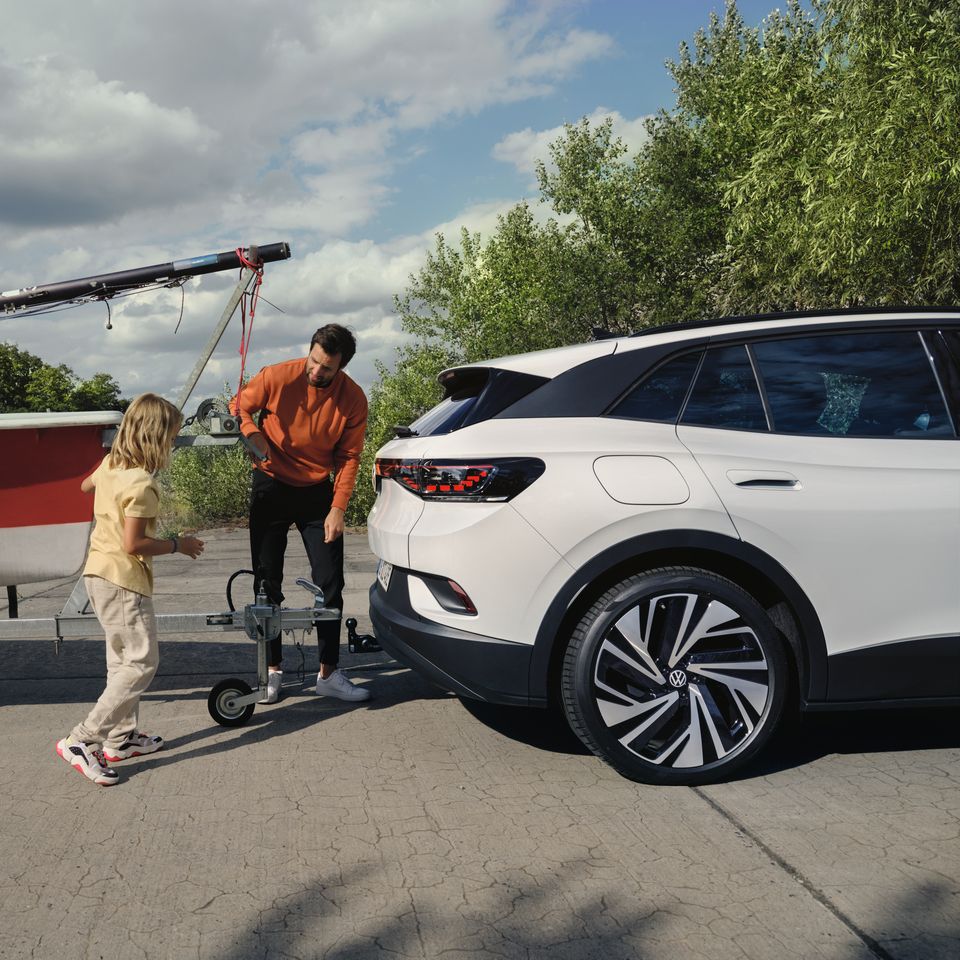 Stranska silhueta belega VW ID.4, moški in deklica na vlečno kljuko priklapljata prikolico s čolnom.