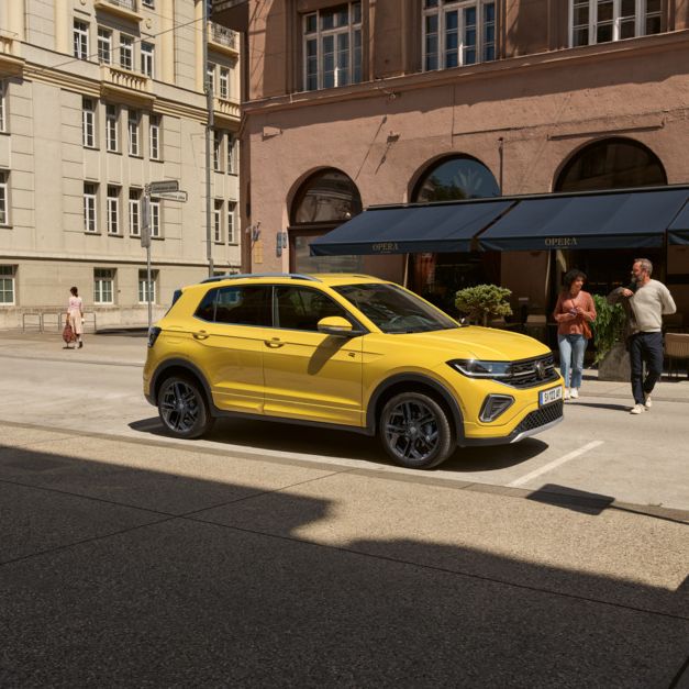 Ein gelber VW T-Cross ist in einer Stadt geparkt, hinter ihm sieht man Menschen