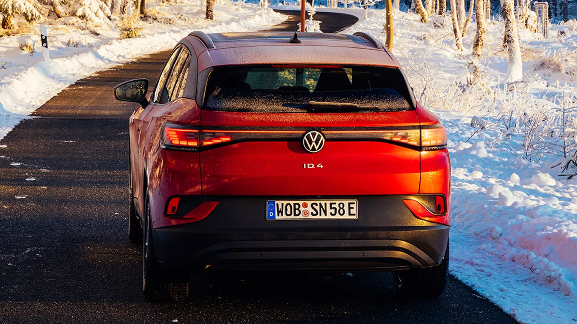 Rdeči Volkswagen ID.4 je parkiran na asfaltni cesti. Okoli je snežna pokrajina z drevesi in rahlo zasneženimi tlemi. Avto ima nemško registrsko tablico WOB-SN58E.