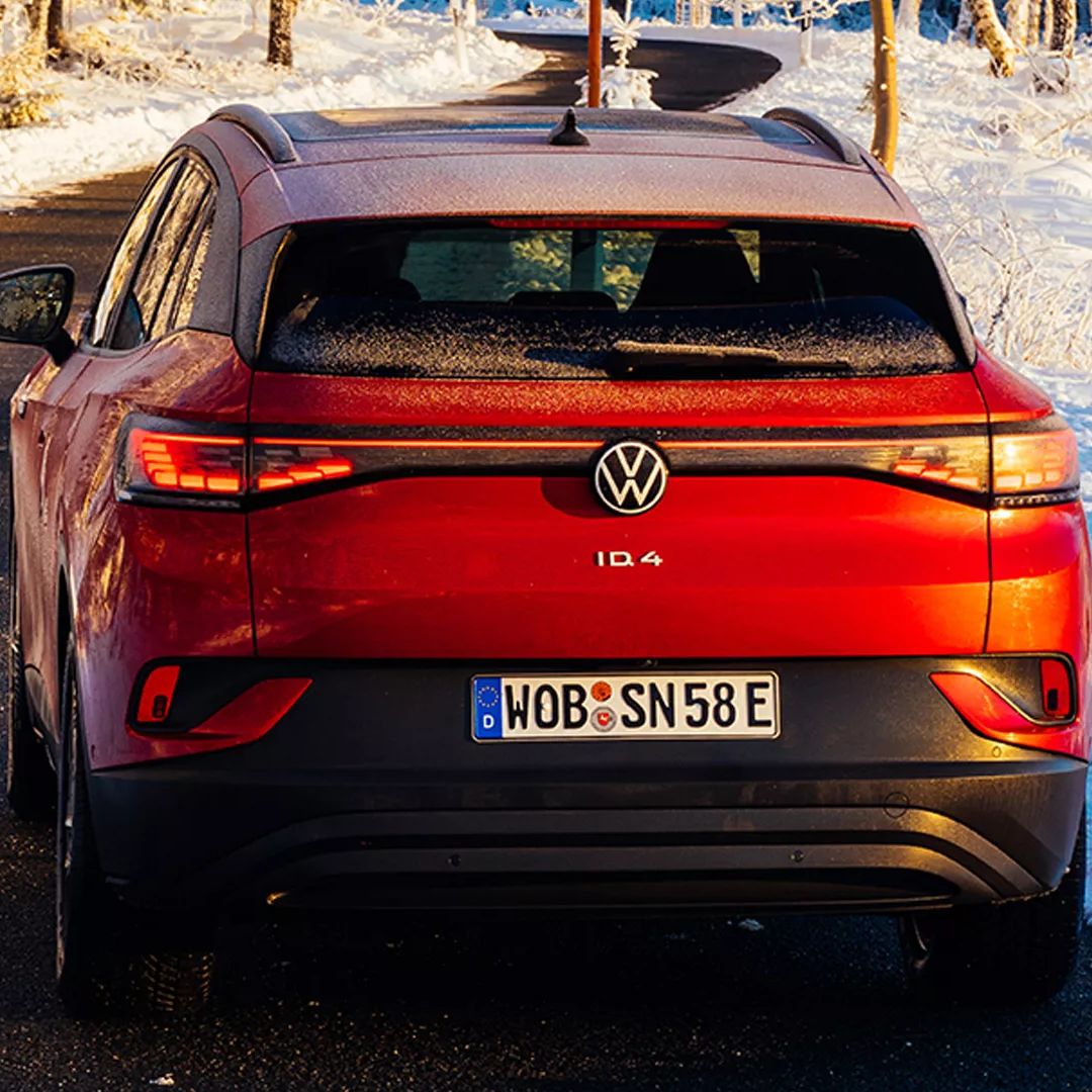 Rdeči Volkswagen ID.4 je parkiran na asfaltni cesti. Okoli je snežna pokrajina z drevesi in rahlo zasneženimi tlemi. Avto ima nemško registrsko tablico WOB-SN58E.