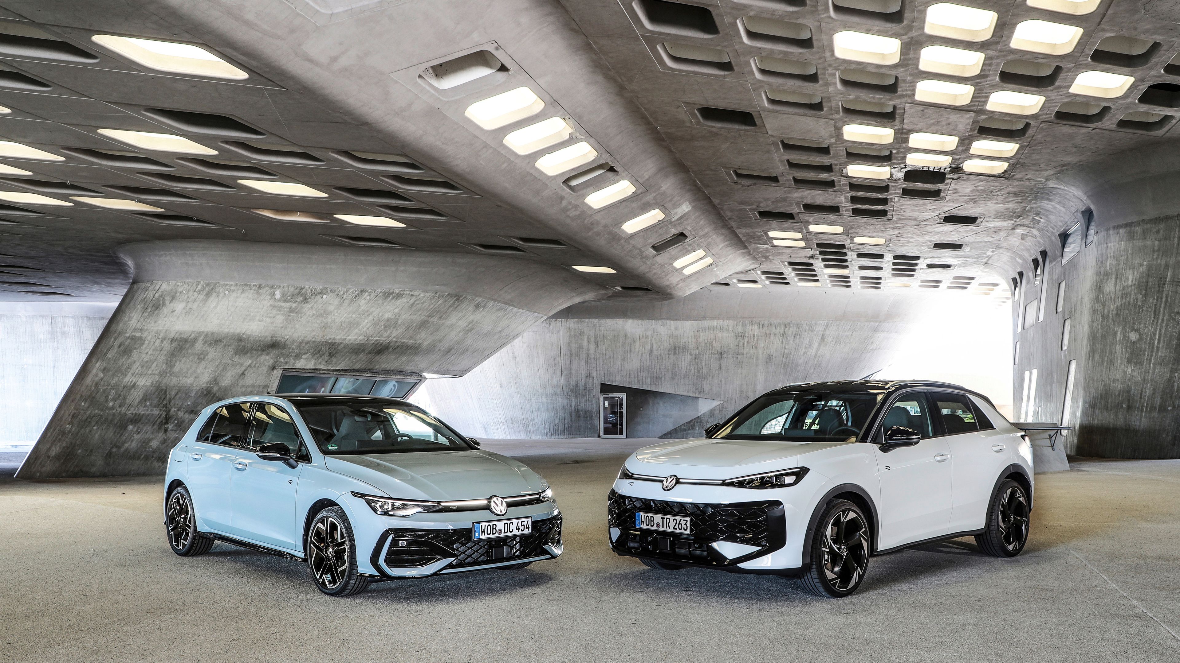 Dva sodobna Volkswagnova avtomobila Golf in T-Roc, svetlo moder Golf in bel T-Roc SUV, sta parkirana drug ob drugem v futuristični betonski garaži z velikimi pravokotnimi odprtinami v stropu. Avtomobila imata športna platišča in poudarjene agresivne linije. 