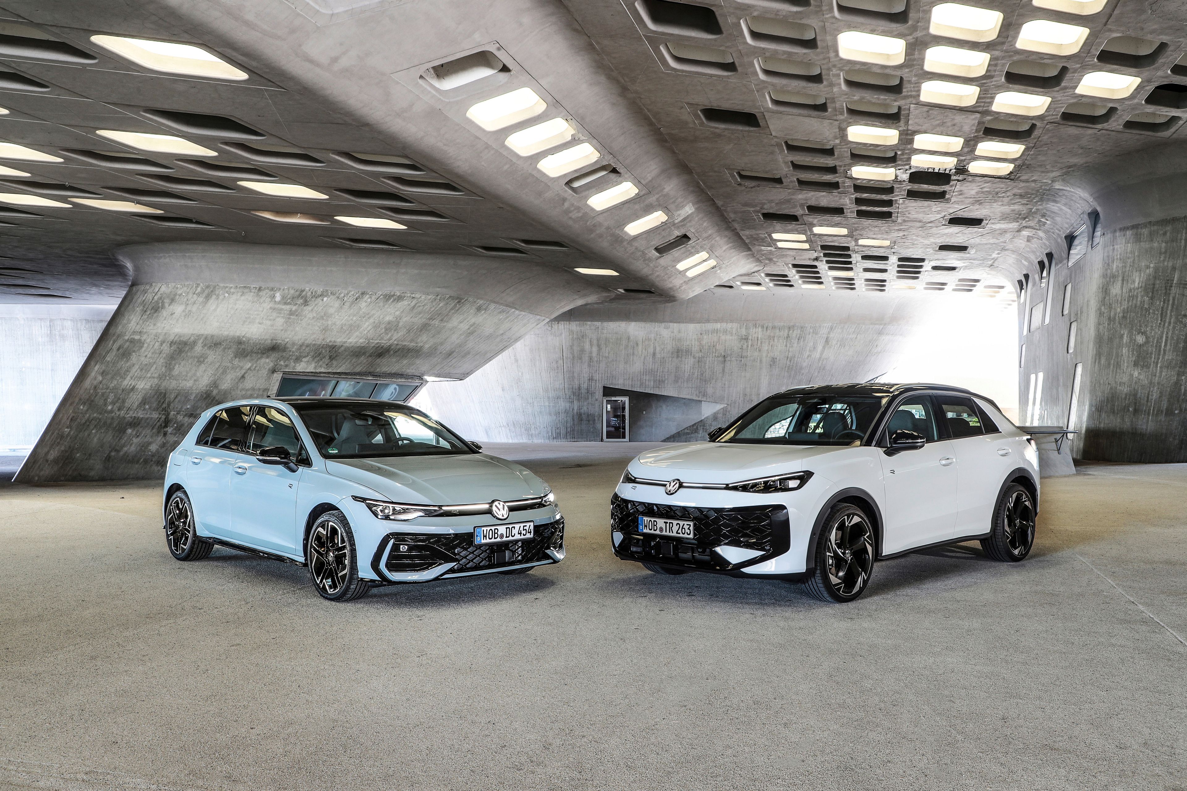 Dva sodobna Volkswagnova avtomobila Golf in T-Roc, svetlo moder Golf in bel T-Roc SUV, sta parkirana drug ob drugem v futuristični betonski garaži z velikimi pravokotnimi odprtinami v stropu. Avtomobila imata športna platišča in poudarjene agresivne linije. 