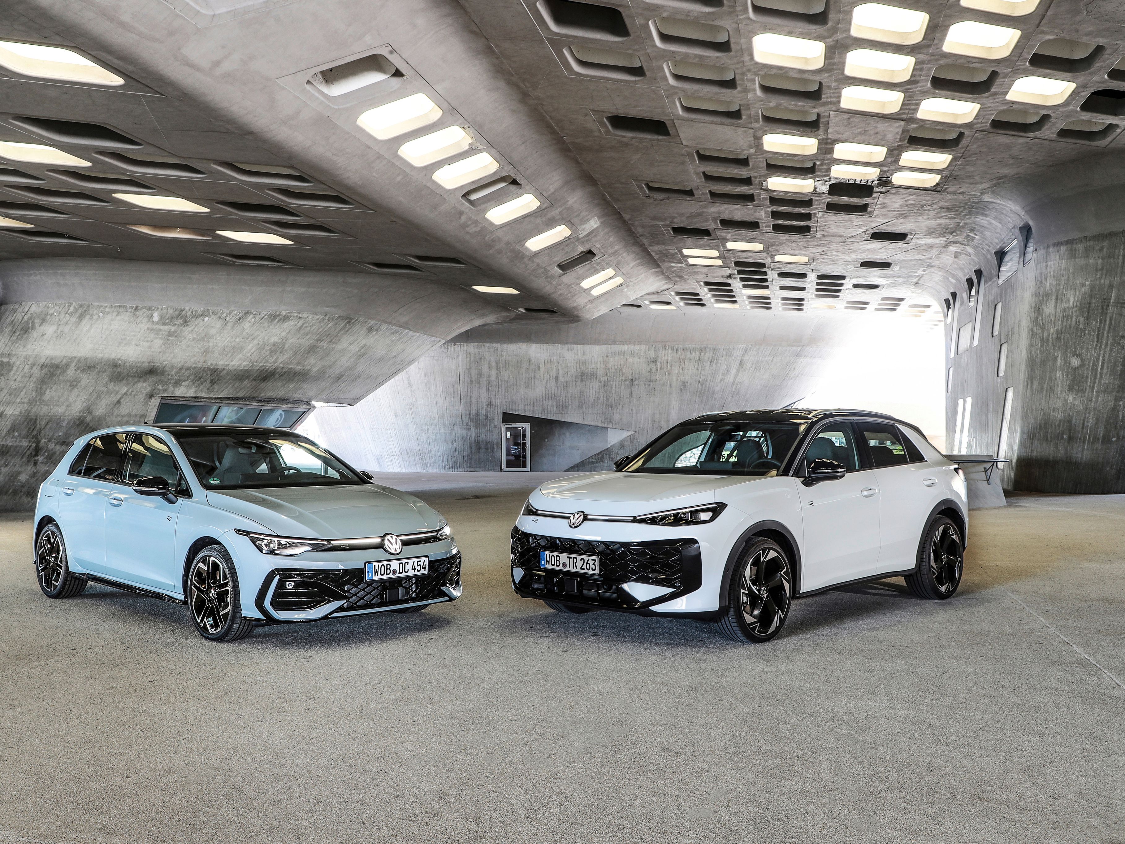 Dva sodobna Volkswagnova avtomobila Golf in T-Roc, svetlo moder Golf in bel T-Roc SUV, sta parkirana drug ob drugem v futuristični betonski garaži z velikimi pravokotnimi odprtinami v stropu. Avtomobila imata športna platišča in poudarjene agresivne linije. 
