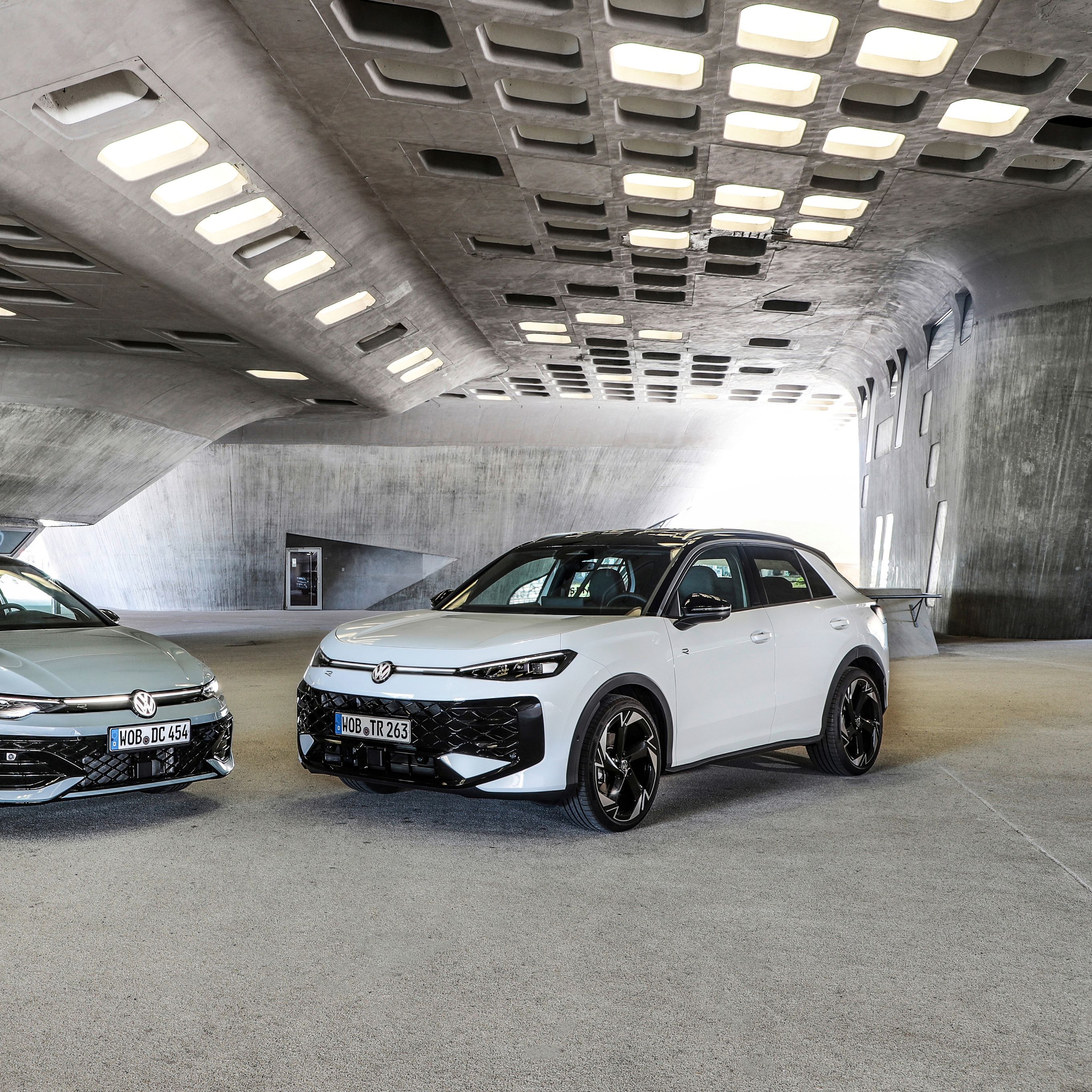 Dva sodobna Volkswagnova avtomobila Golf in T-Roc, svetlo moder Golf in bel T-Roc SUV, sta parkirana drug ob drugem v futuristični betonski garaži z velikimi pravokotnimi odprtinami v stropu. Avtomobila imata športna platišča in poudarjene agresivne linije. 