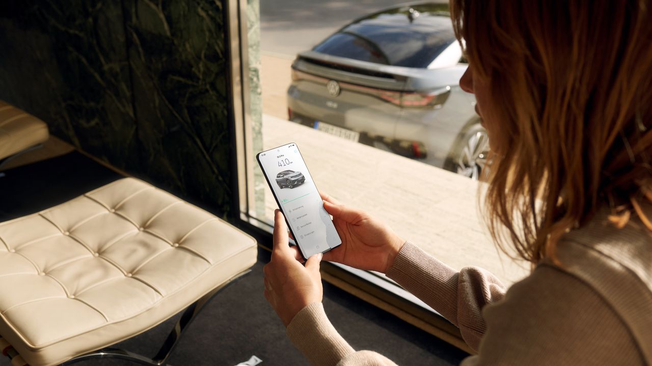 Eine Frau hält ein Smartphone in der Hand mit der We Connect App. Diese liefert Daten zu einem ID.5, der vor dem Fenster geparkt ist. 