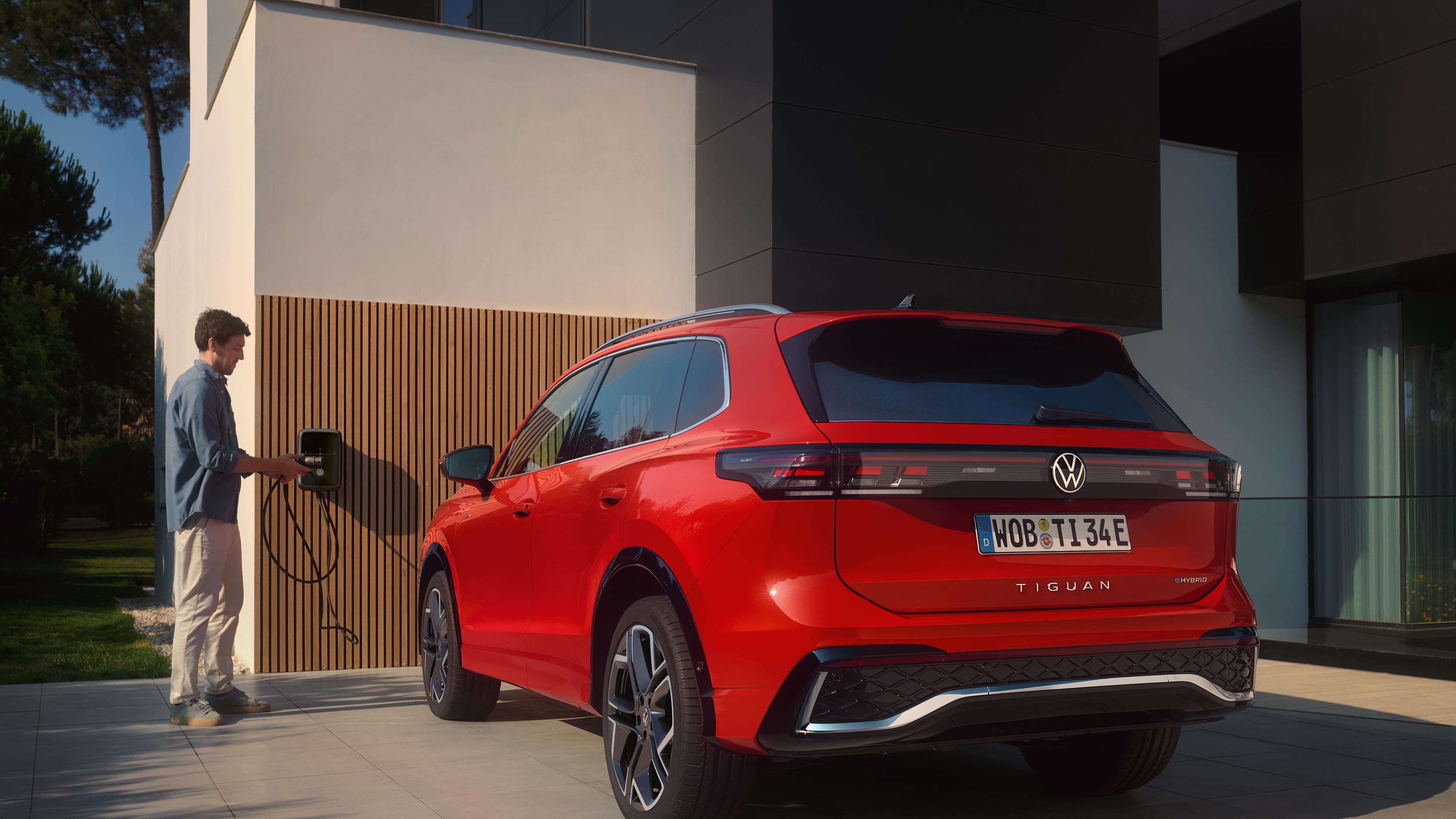 Rdeč VW Tiguan eHybrid je parkiran pred hišo, moški ga polni prek stenske polnilne postaje na hiši.