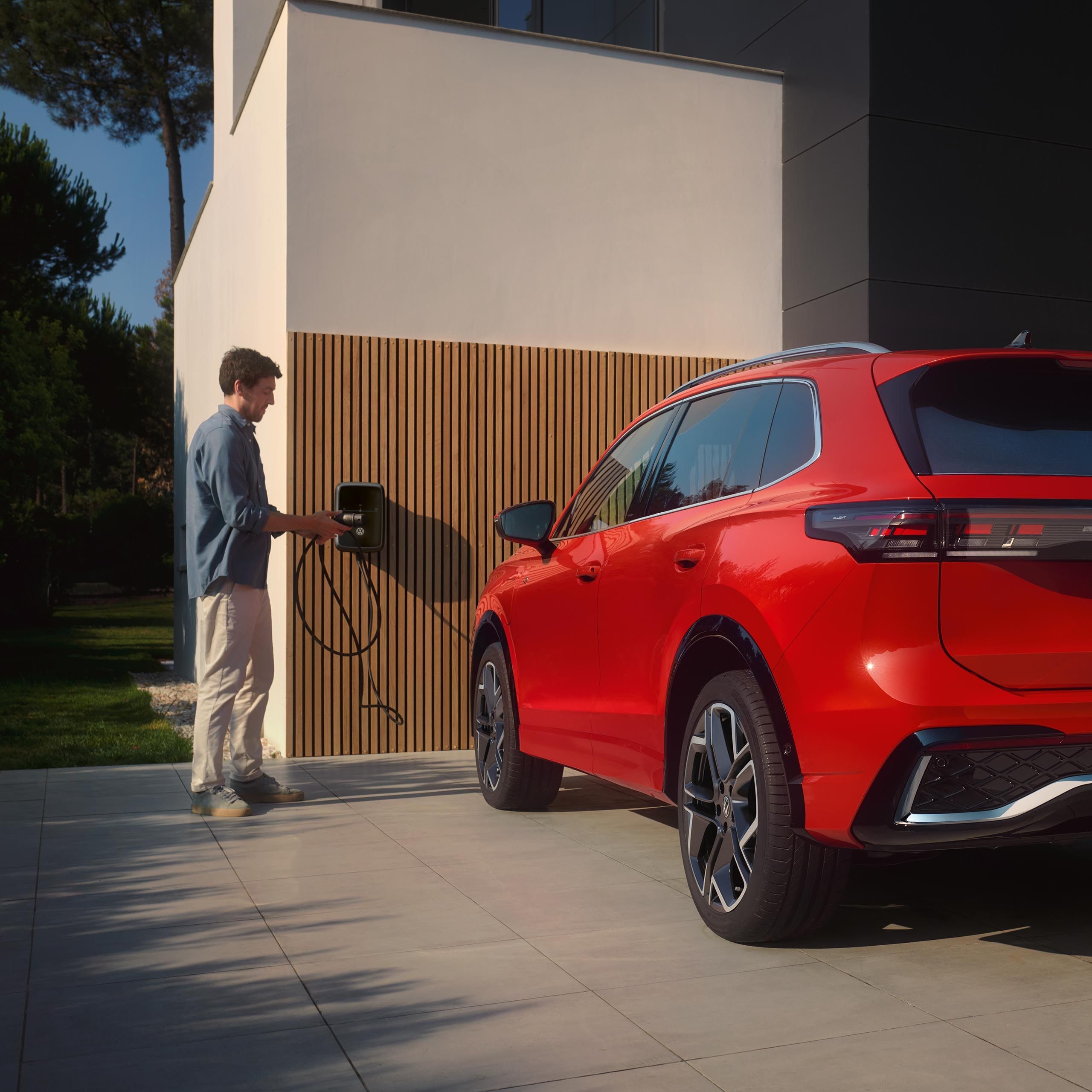 Rdeč VW Tiguan eHybrid je parkiran pred hišo, moški ga polni prek stenske polnilne postaje na hiši.