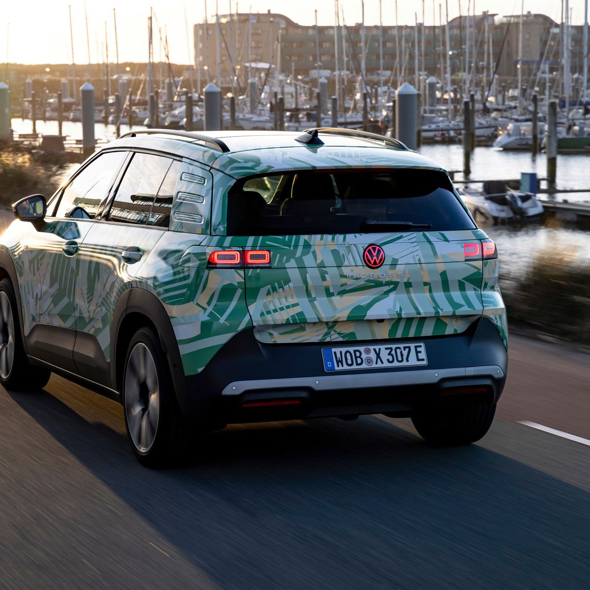 Električni avtomobil Volkswagen SUV vozi po cesti ob pristanišču ob sončnem zahodu. V ozadju je mogoče videti številne jadrnice in jahte. Cesta in podeželje se zdita mirna in spokojna.