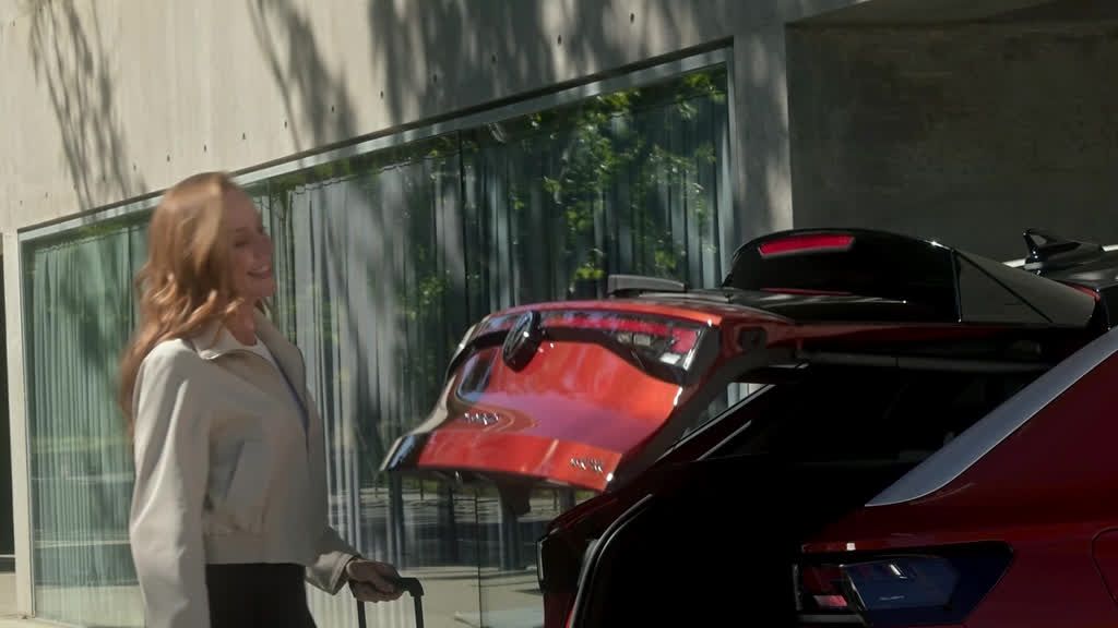 Ženska s kovčkom stoji poleg parkiranega avtomobila rdeče barve z odprtim prtljažnikom. 