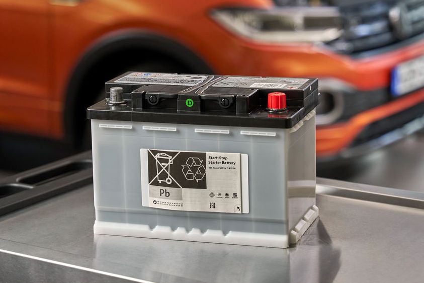 Akumulator za avto je postavljen na vozičku v mehanični delavnici. V ozadju je oranžen T-Cross z odprtim pokrovom motorja.