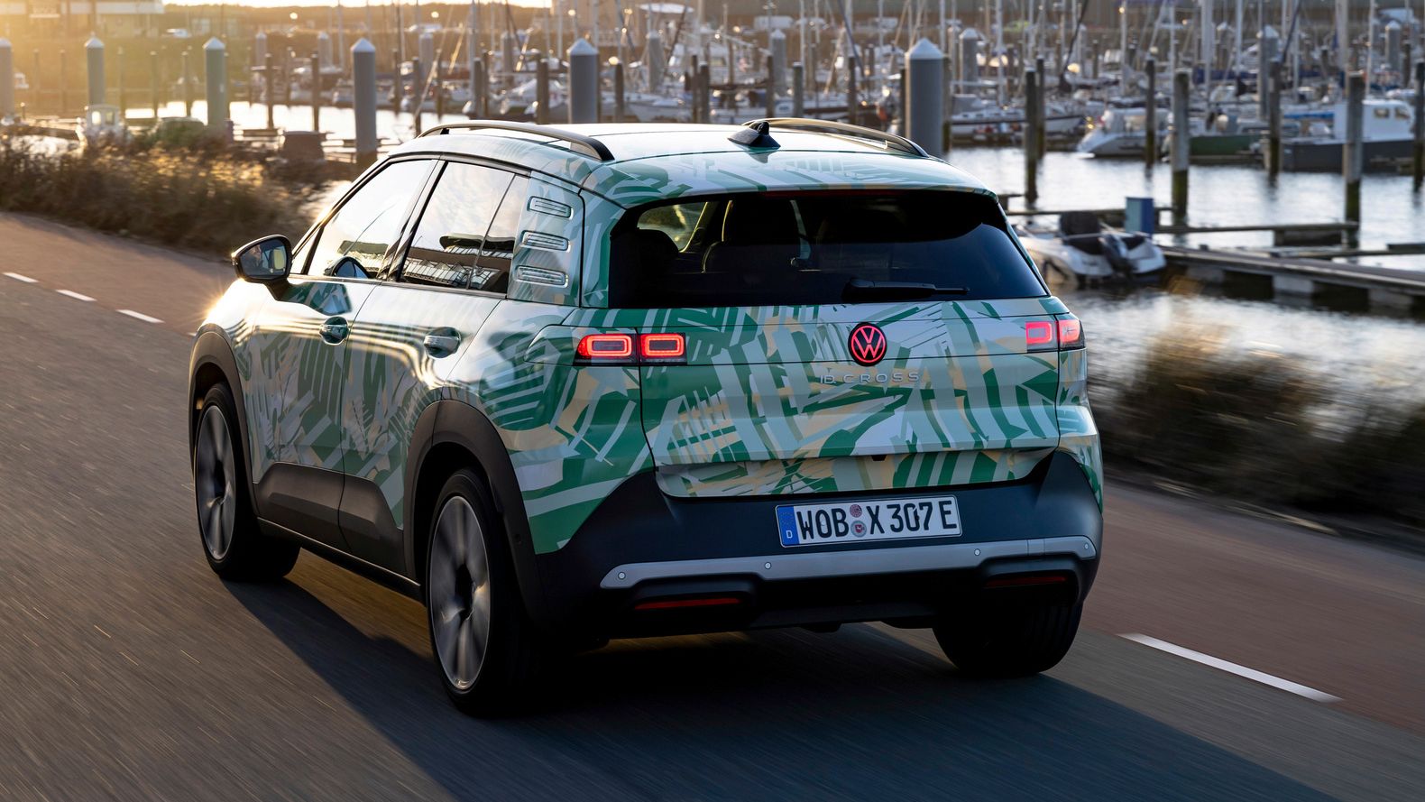 Električni avtomobil Volkswagen SUV vozi po cesti ob pristanišču ob sončnem zahodu. V ozadju je mogoče videti številne jadrnice in jahte. Cesta in podeželje se zdita mirna in spokojna.