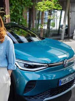 Ženska s pametnim telefonom v roki stoji pred modrim VW ID.4. V ozadju je vidna stenska polnilna postaja, na katero je priključeno vozilo.