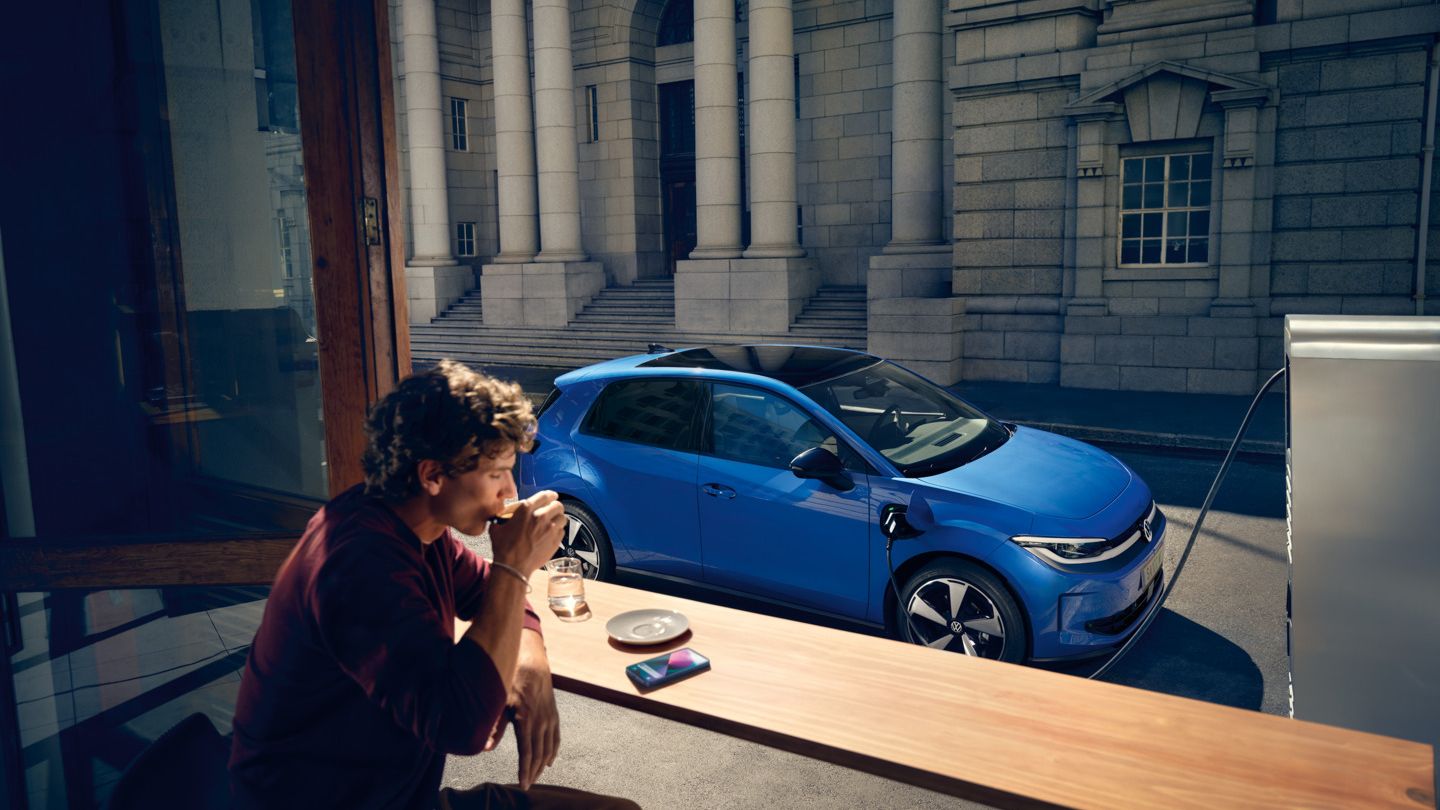 Moški sedi v kavarni, skozi odprto okno je viden modri VW ID. Polo Style, ki polni na polnilni postaji.