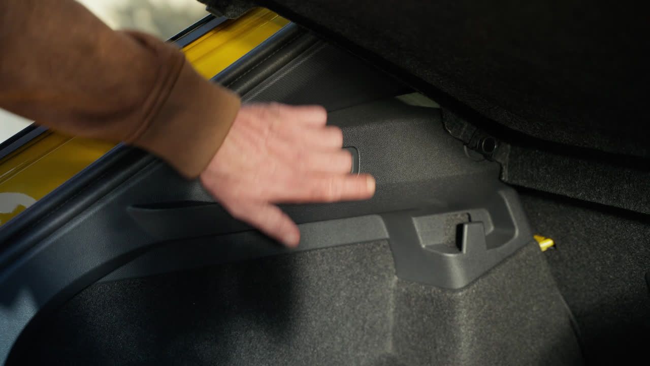 Roka v rjavi jakni se dotika notranjosti prtljažnika avtomobila. Prtljažnik je odprt, viden je del rumenega avtomobila. Notranjost prtljažnika je sestavljena iz črnih in sivih delov.