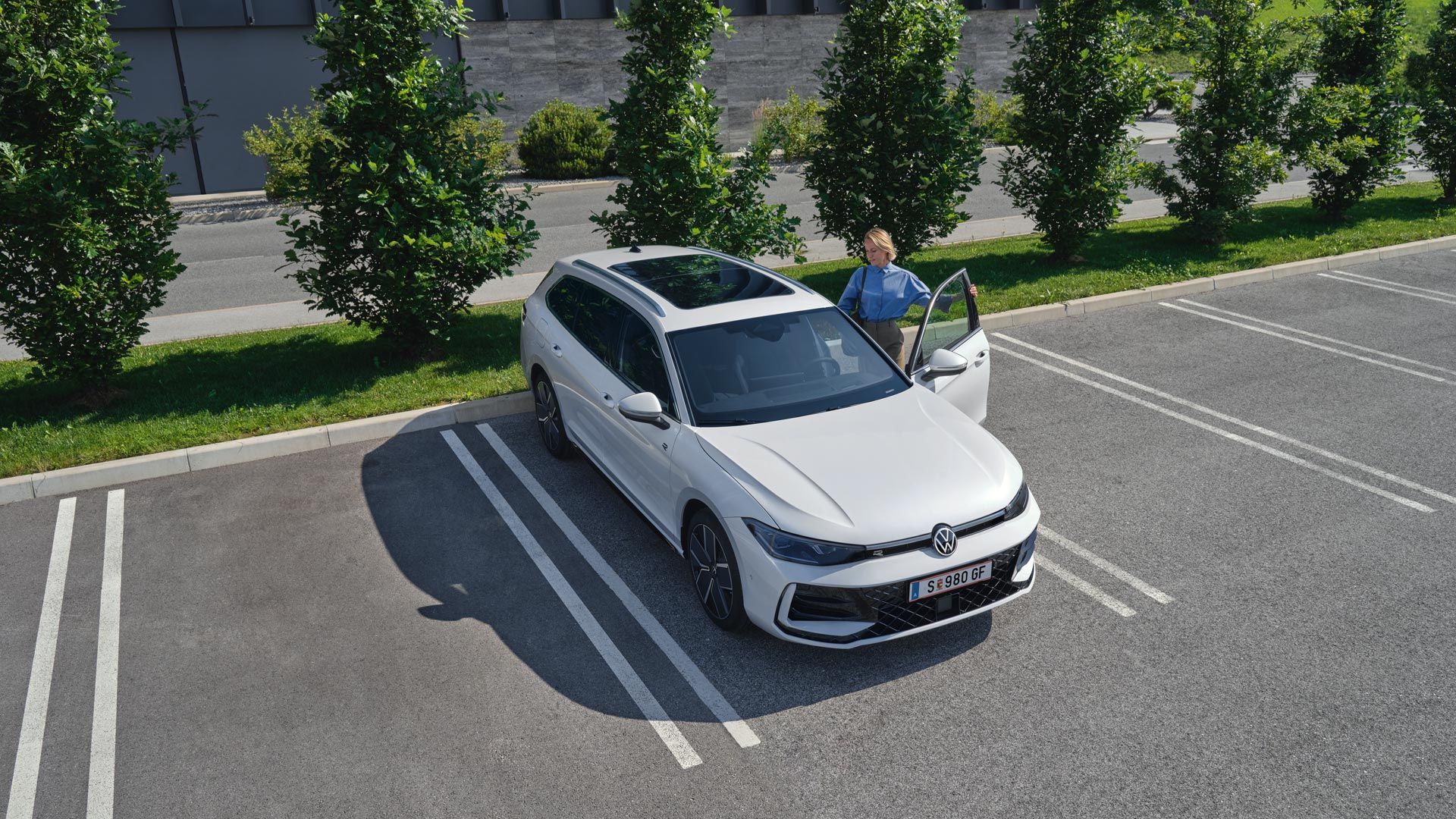 Pogled od zgoraj na bel VW Passat na parkirišču, ženska vstopa na voznikovi strani.