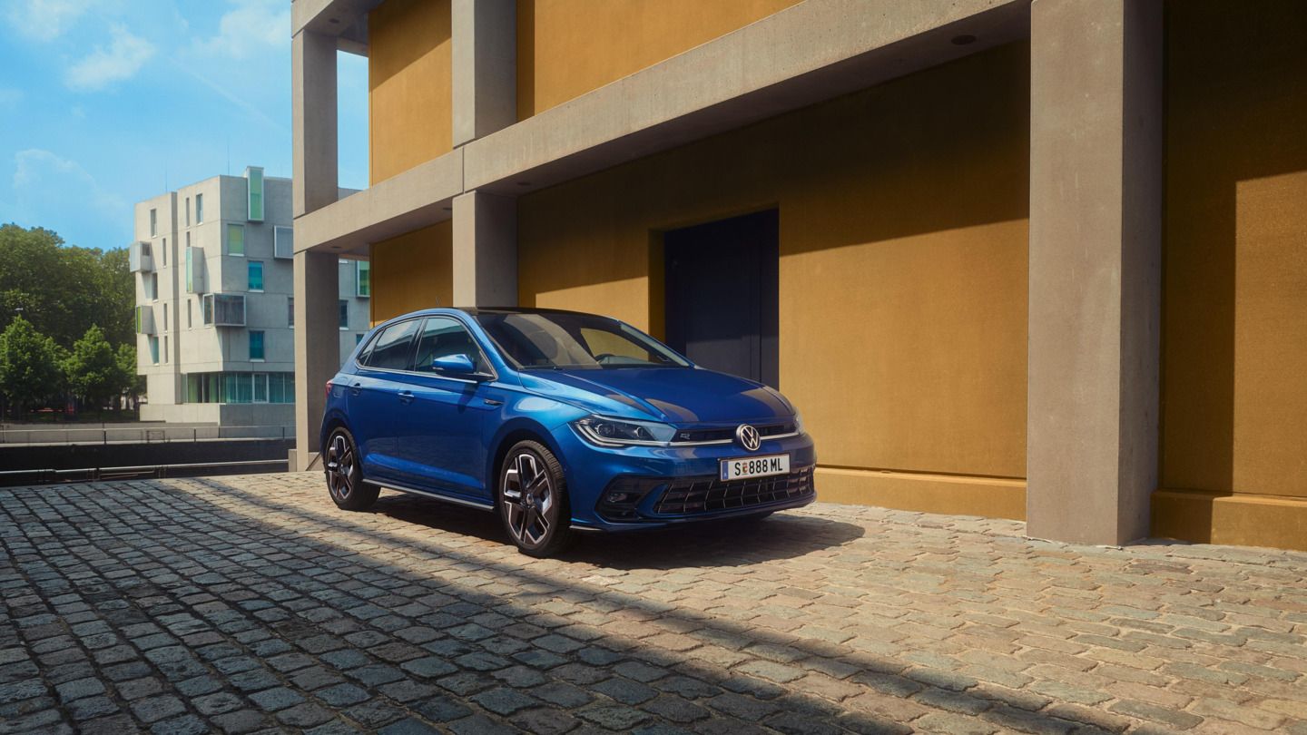 Moder Polo R-Line je parkiran pred stavbo na tlakovani cesti. Prikazana sta sprednji del in sovoznikova stran. 