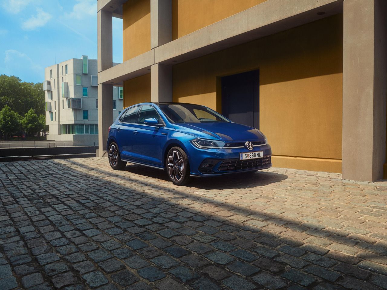 Moder Polo R-Line je parkiran pred stavbo na tlakovani cesti. Prikazana sta sprednji del in sovoznikova stran. 