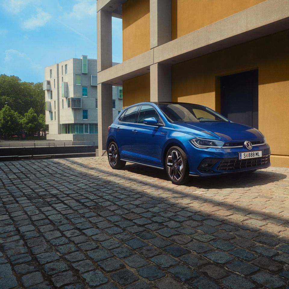 Moder Polo R-Line je parkiran pred stavbo na tlakovani cesti. Prikazana sta sprednji del in sovoznikova stran. 