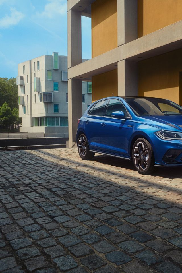Moder Polo R-Line je parkiran pred stavbo na tlakovani cesti. Prikazana sta sprednji del in sovoznikova stran. 