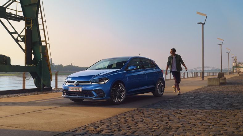 Moder VW Polo je parkiran v pristanišču, od strani je prikazan sprednji del. Moški gre mimo zadnjega dela avtomobila. 