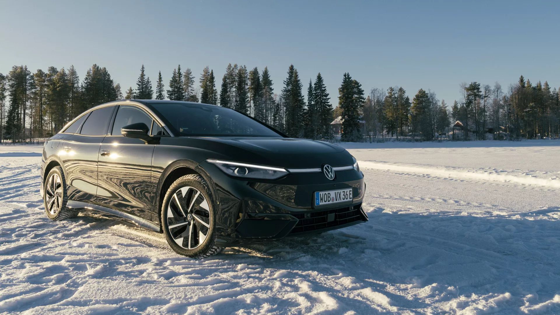 Črn Volkswagen ID.7 stoji na zasneženi pokrajini pod jasnim modrim nebom. V ozadju je vrstica zasneženih dreves in nekaj odmaknjenih hiš. 