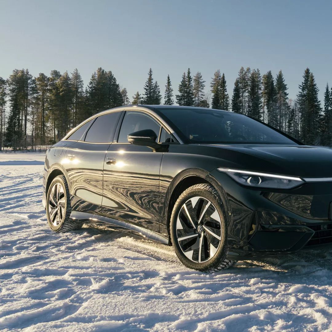 Črn Volkswagen ID.7 stoji na zasneženi pokrajini pod jasnim modrim nebom. V ozadju je vrstica zasneženih dreves in nekaj odmaknjenih hiš. 