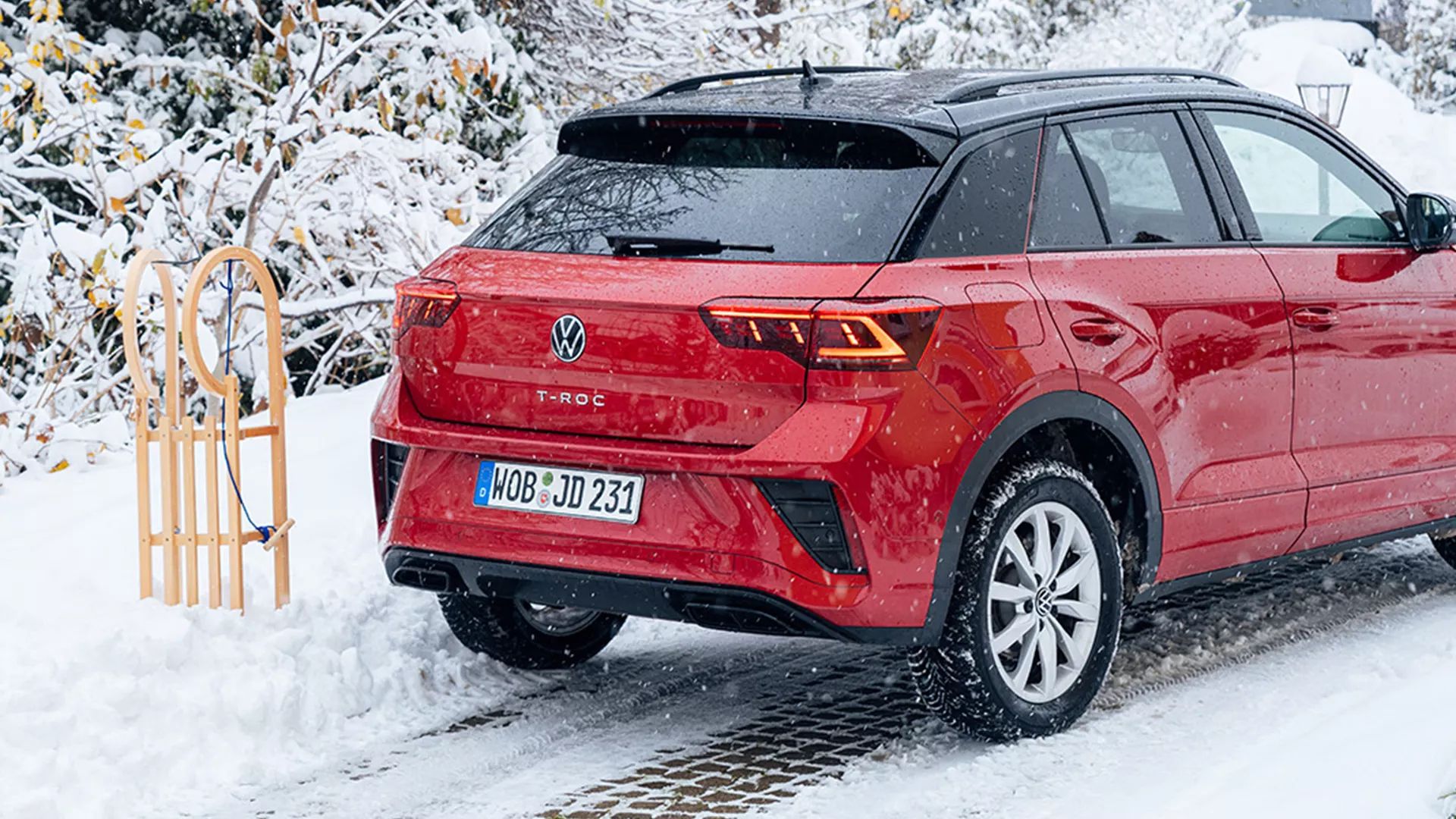Rdeč Volkswagen T-Roc je parkiran na zasneženi cesti ob lesenih sankah. V ozadju so zasnežena drevesa in grmičevje. Na tleh so vidne stopinje v snegu.
