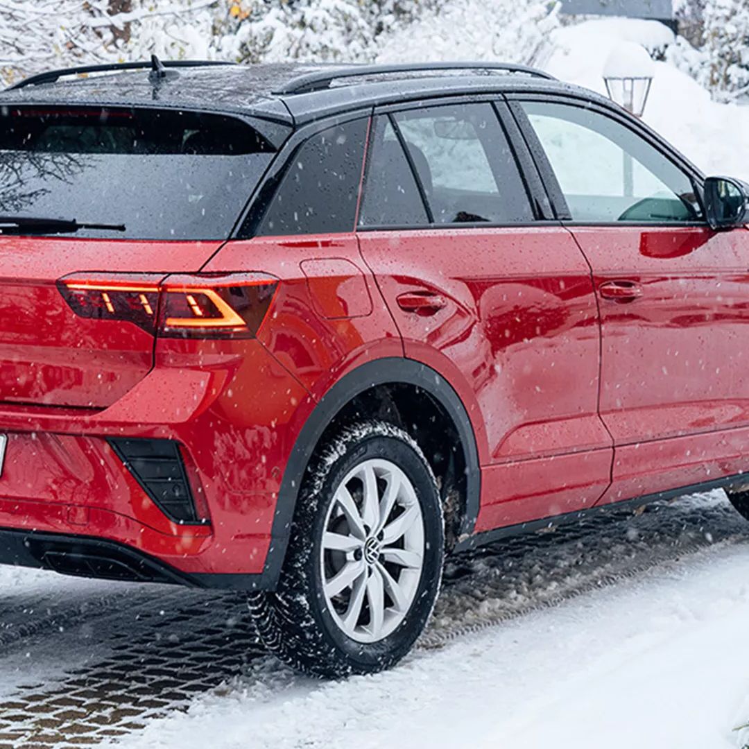 Rdeč Volkswagen T-Roc je parkiran na zasneženi cesti ob lesenih sankah. V ozadju so zasnežena drevesa in grmičevje. Na tleh so vidne stopinje v snegu.