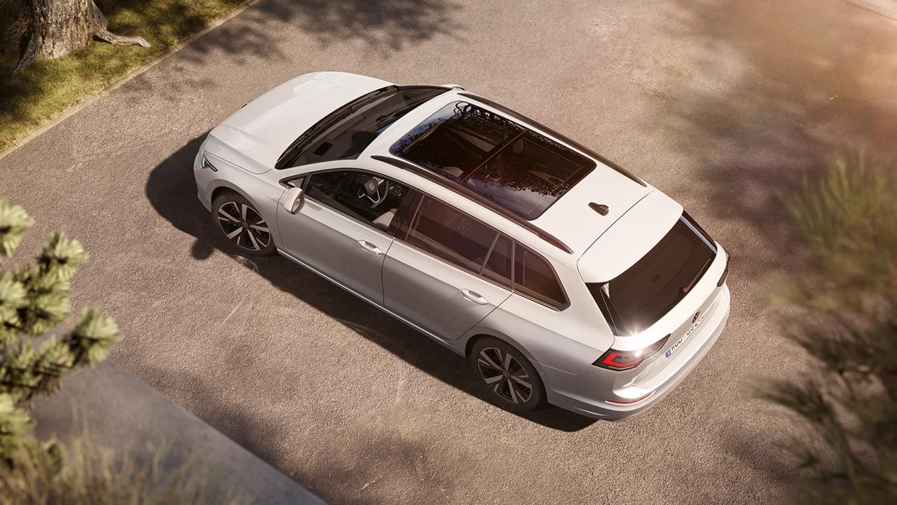 Srebrn VW Golf Variant je parkiran na dovozni poti, fotografiran od zgoraj pod kotom. Avto ima panoramsko strešno okno, elegantne linije in sodoben dizajn platišč. V ozadju so mehke sence dreves in topel sončni sij.