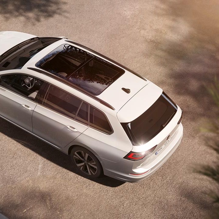 Srebrn VW Golf Variant je parkiran na dovozni poti, fotografiran od zgoraj pod kotom. Avto ima panoramsko strešno okno, elegantne linije in sodoben dizajn platišč. V ozadju so mehke sence dreves in topel sončni sij.