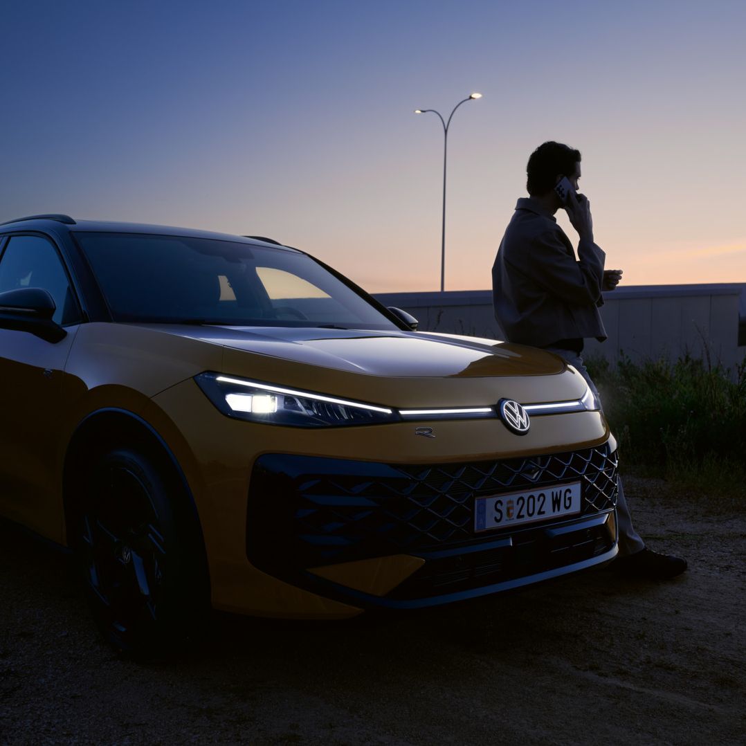 Moški stoji poleg VW T-Roc R-Line z prižganimi žarometi in osvetljenim logotipom.
