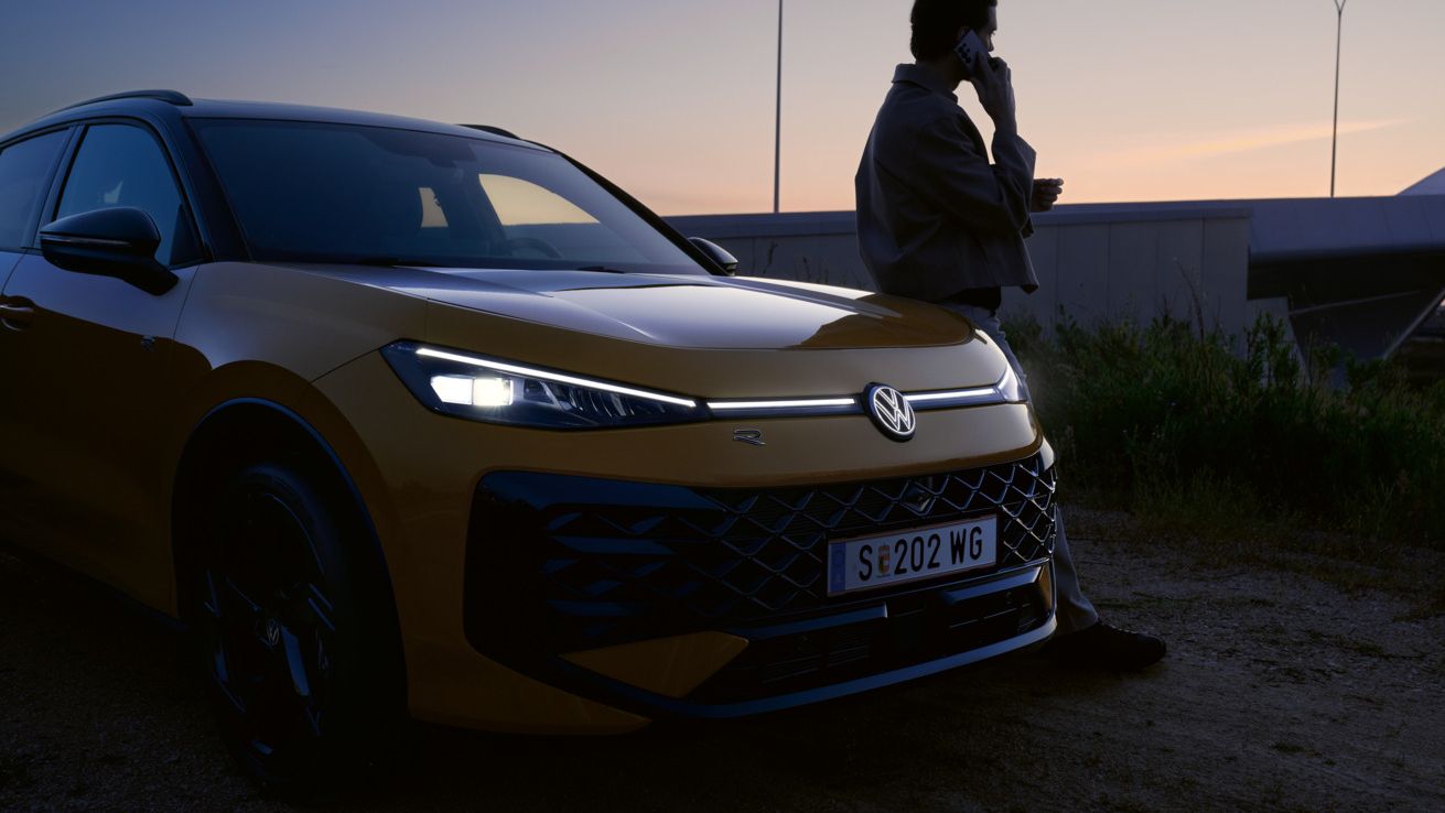 Moški stoji poleg VW T-Roc R-Line z prižganimi žarometi in osvetljenim logotipom.