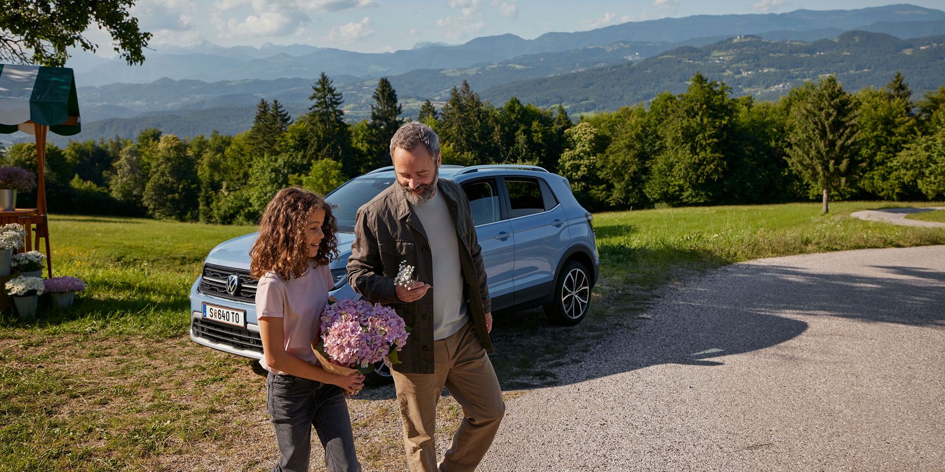 Modri VW T-Cross je parkiran na travniku v gorah. Pred njim se dekle in moški sprehajata s cvetjem v rokah.
