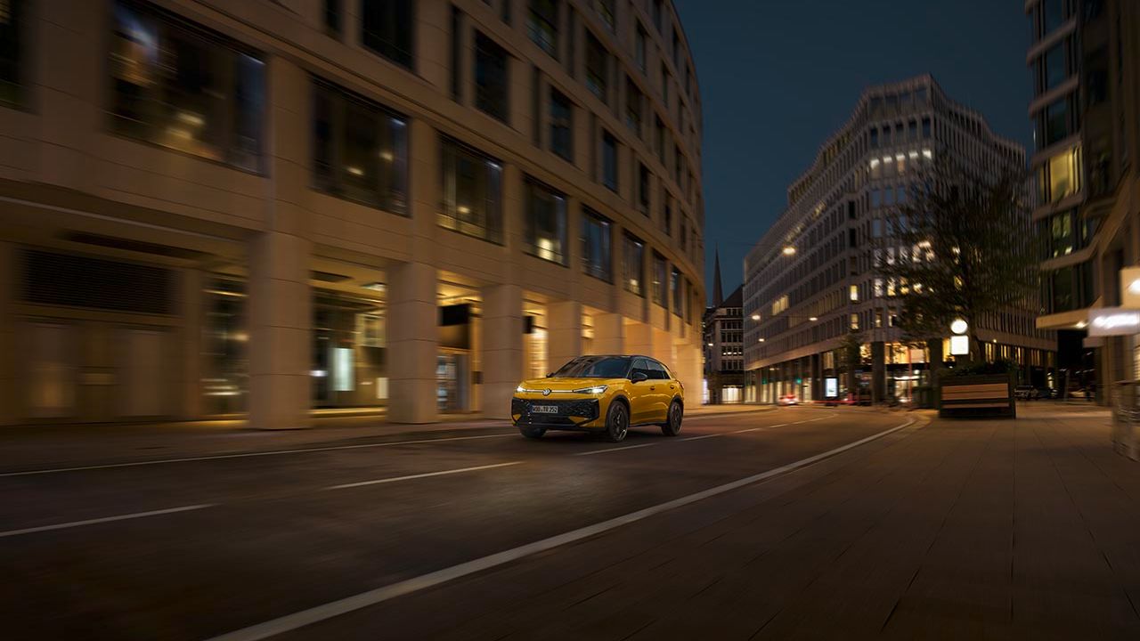 Rumeni Volkswagen T-Roc vozi po prazni mestni ulici ponoči. Okoli avtomobila so vidne moderne stavbe z osvetljenimi okni. Ulica je mirna brez pešcev.