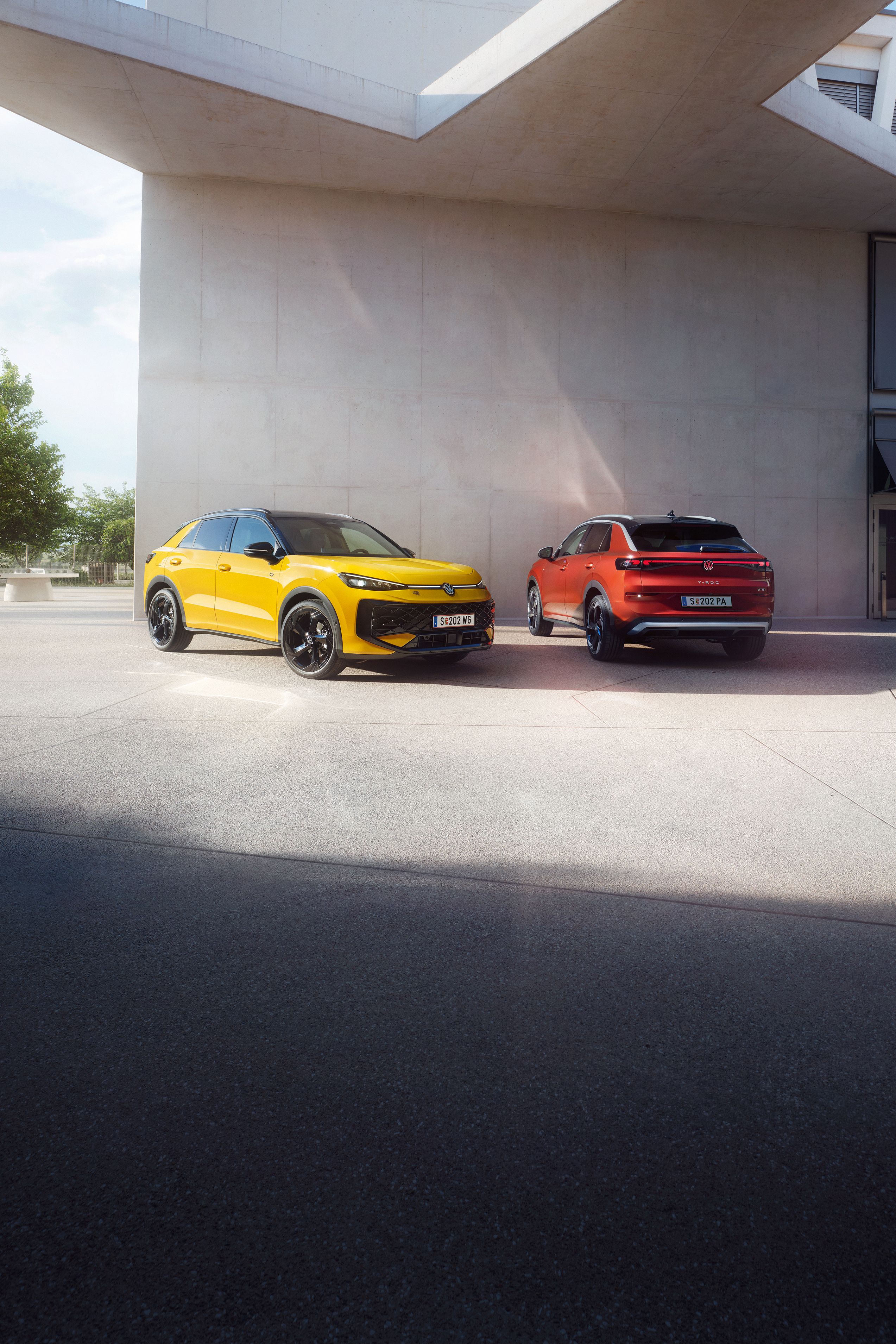 Rumeni VW T-Roc R-Line in rdeči VW T-Roc Style stojita drug ob drugem pred sodobno stavbo.